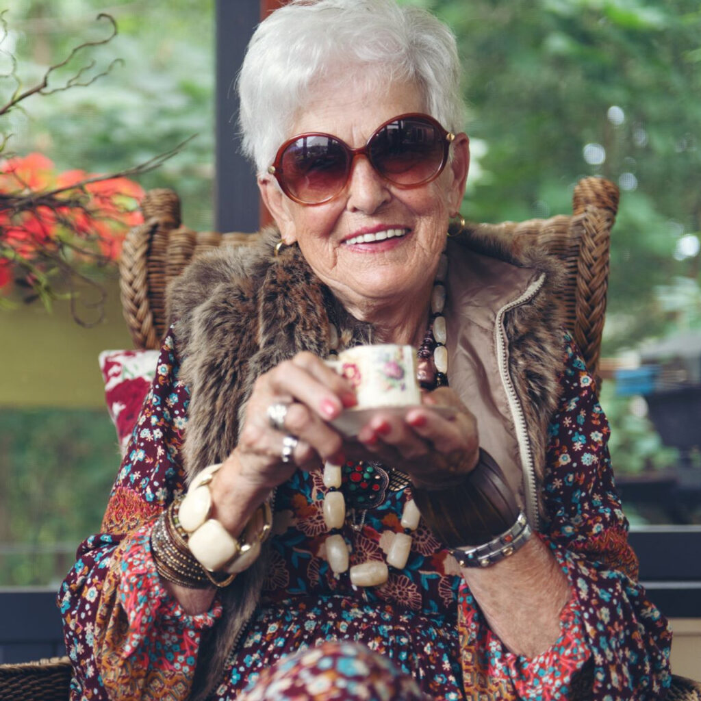 Äldre kvinna med mörka solglasögon dricker en kopp kaffe på sin uteplats/balkong.