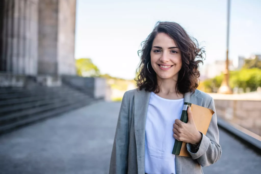 En glad kvinna med mörkt hår , iklädd vit tröja och en grå kavaj, håller i ett anteckningsblock nedanför en stentrapp som leder upp till en domstolsbyggnad.