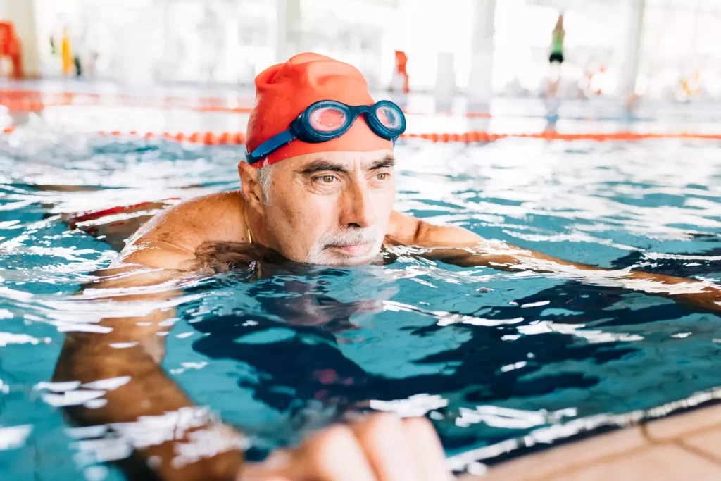 Äldre man med orange badmössa och simglasögon vid kanten av en pool i ett badhus